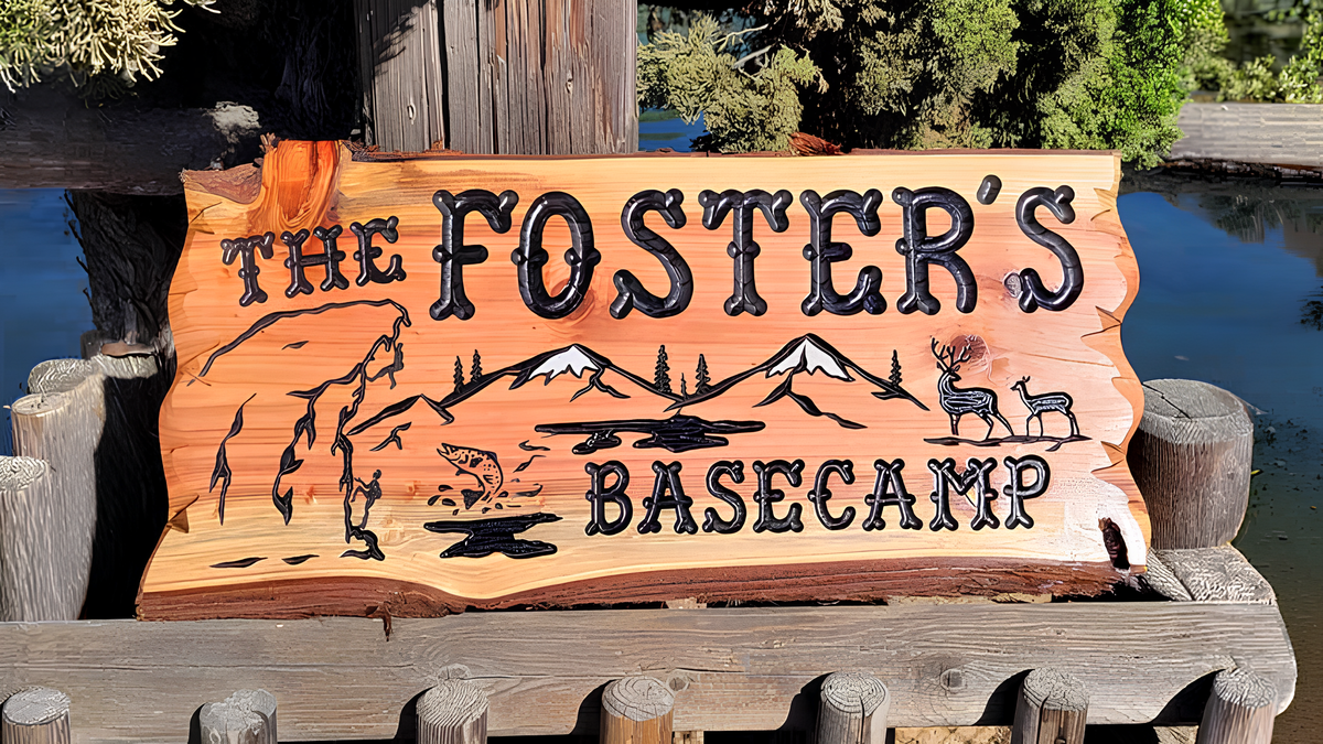 A rustic hand-carved wooden sign reading 'THE FOSTER'S BASECAMP' with mountain, deer, and campfire illustrations, displayed outdoors on a wooden dock or railing near water and trees.