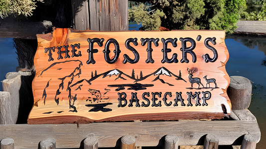 A rustic hand-carved wooden sign reading 'THE FOSTER'S BASECAMP' with mountain, deer, and campfire illustrations, displayed outdoors on a wooden dock or railing near water and trees.